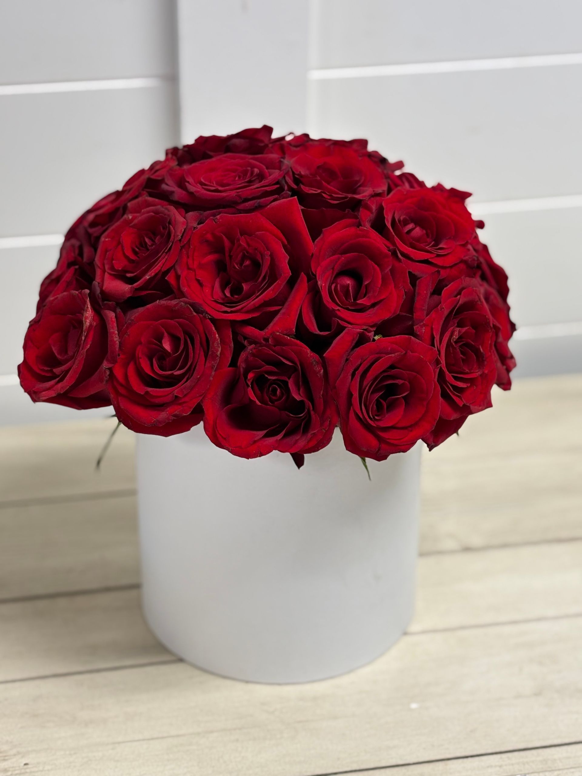 A dense bouquet of deep red roses arranged in a simple white cylindrical vase against a white paneled wall.