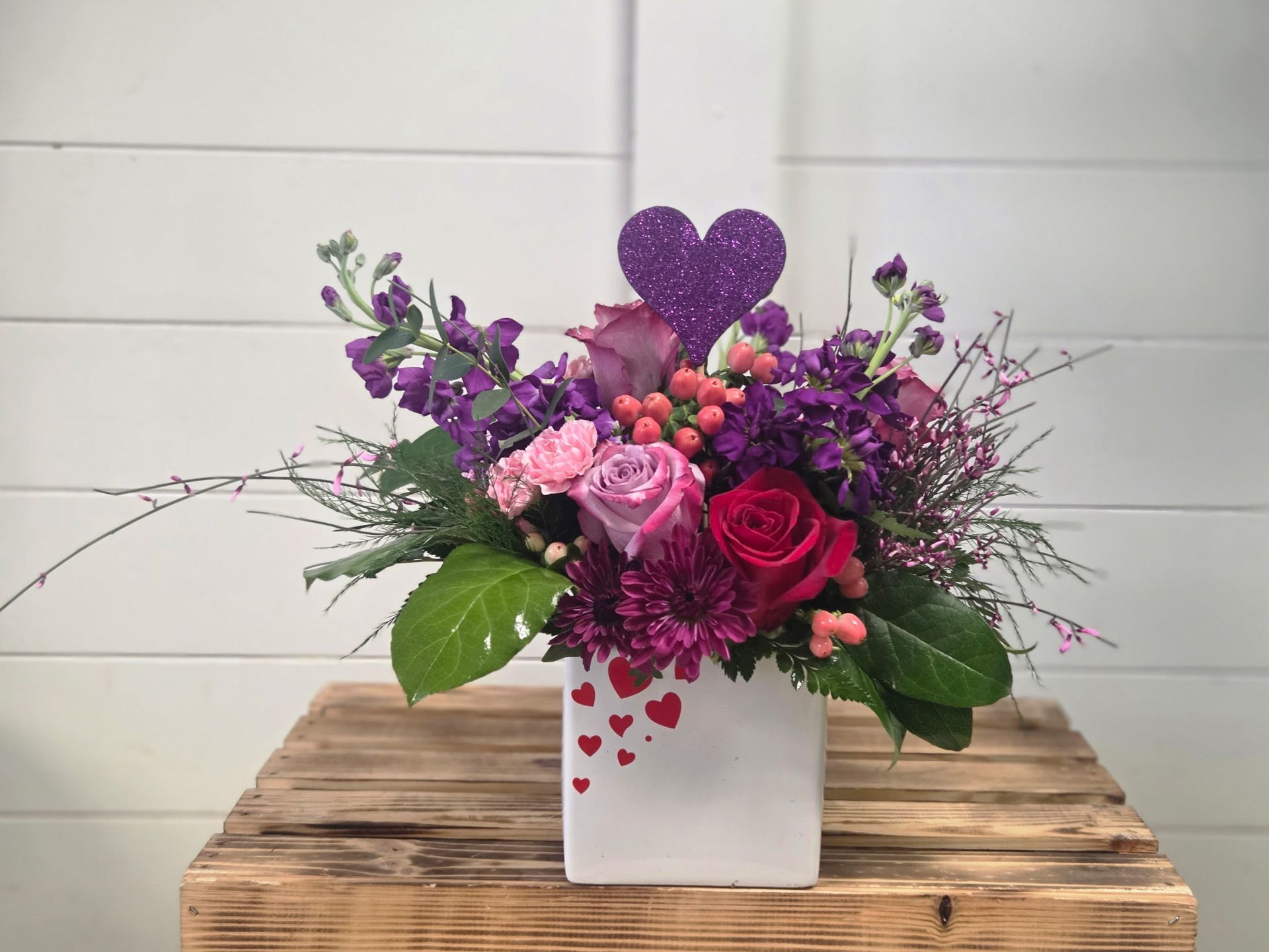 A floral arrangement of purple and pink roses and stock in a white vase adorned with small red hearts and a purple heart.