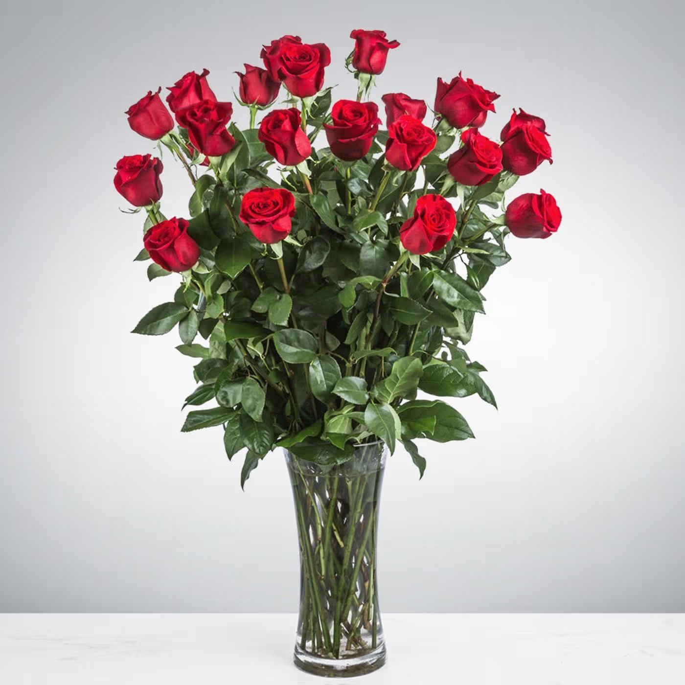 A large, lush bouquet of bright red roses arranged in a clear glass vase against a plain grey background.