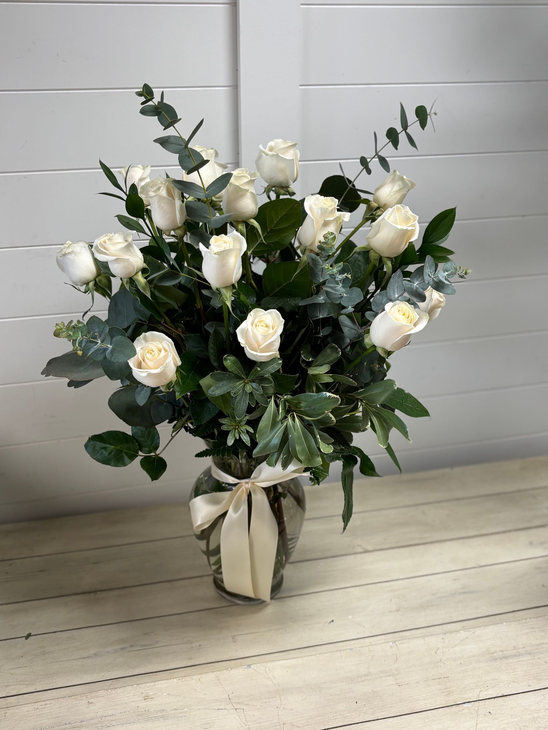 A bouquet of white roses and green eucalyptus leaves in a clear glass vase with a white ribbon, set against a white wall.
