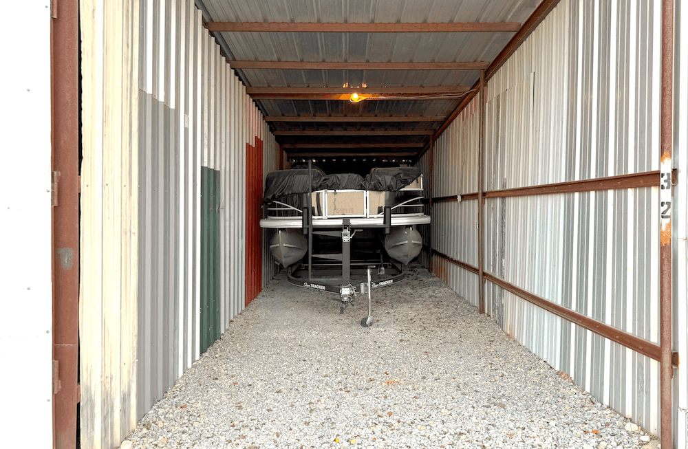 Boat on trailer in gravel-floored storage unit with corrugated metal walls and ceiling.