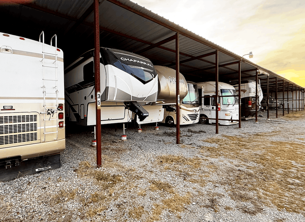 Row of RVs parked under a metal-roofed shelter in a gravel lot; dusk lighting.