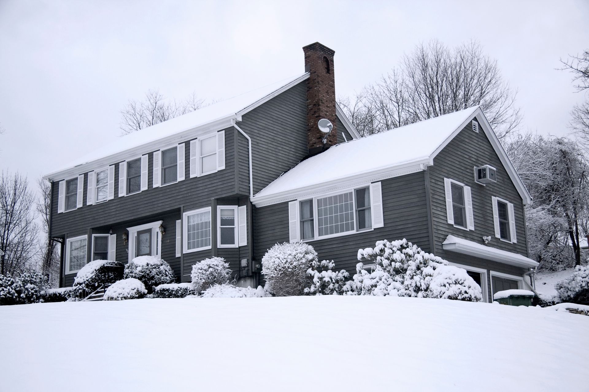 Two-story gray house covered in snow with white shutters, bushes, and trees on a snowy hillside.