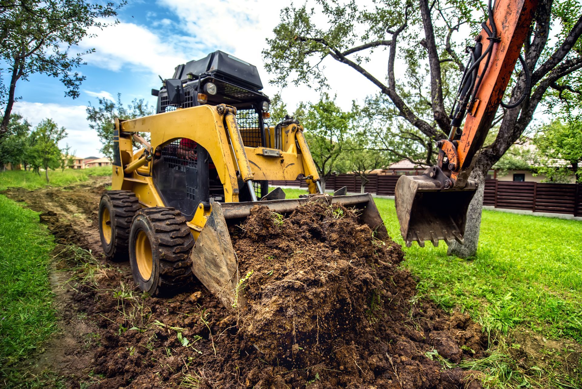 Yellow skid steer and orange excavator digging in a grassy yard.