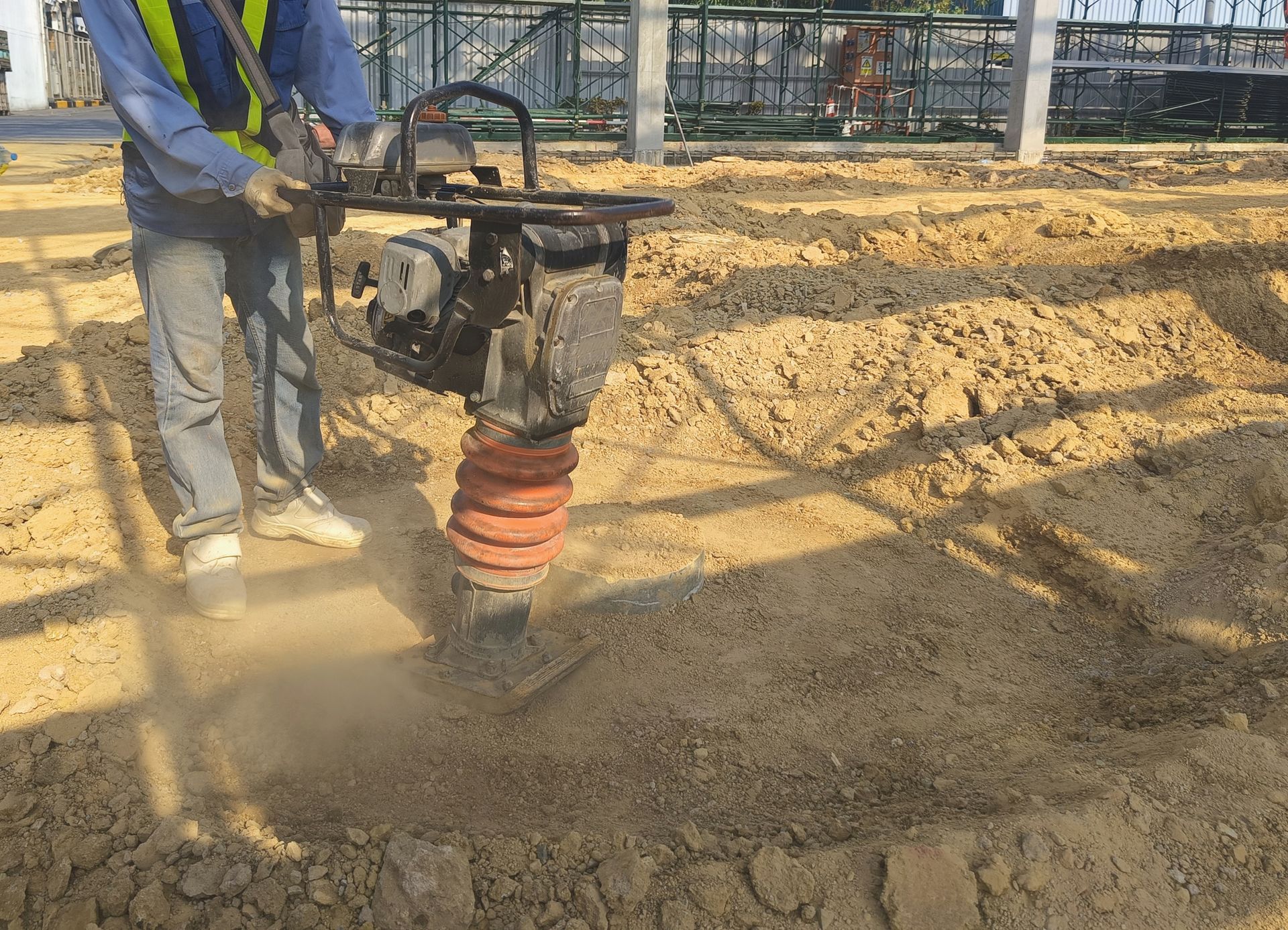 Worker operating a vibrating plate compactor on a dirt surface at a construction site