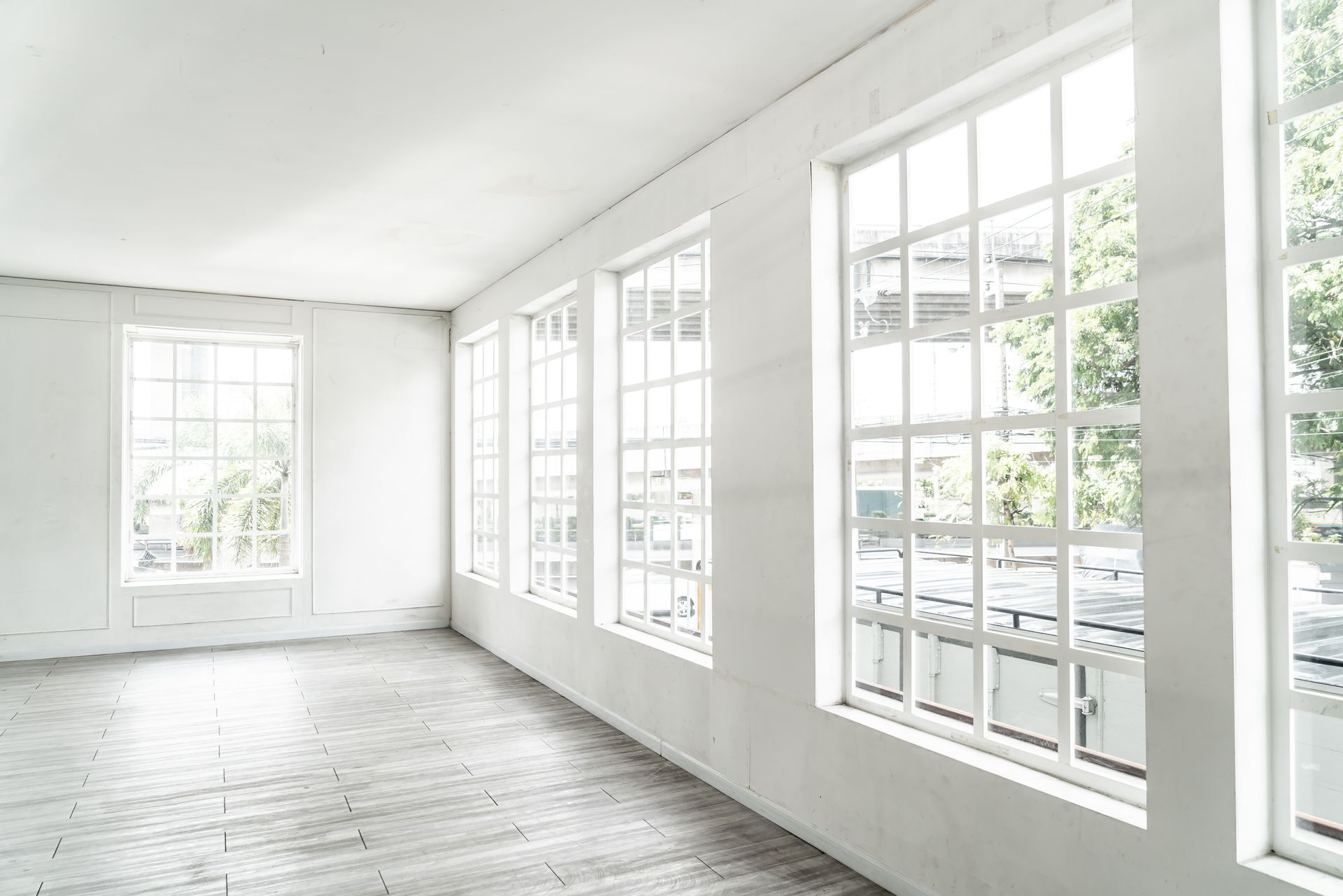 A bright, minimalist room featuring multiple tall, white-framed grid windows along a wall with light-colored wood flooring.