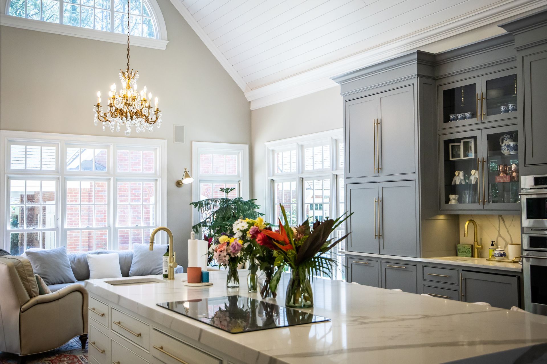 A kitchen with a large marble island, grey cabinetry, a crystal chandelier, and large windows with seating.
