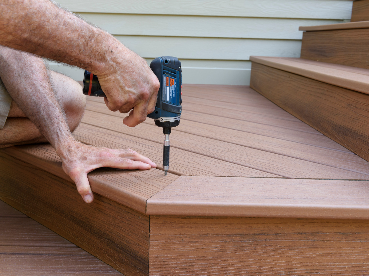 A man is using a drill on a wooden deck.