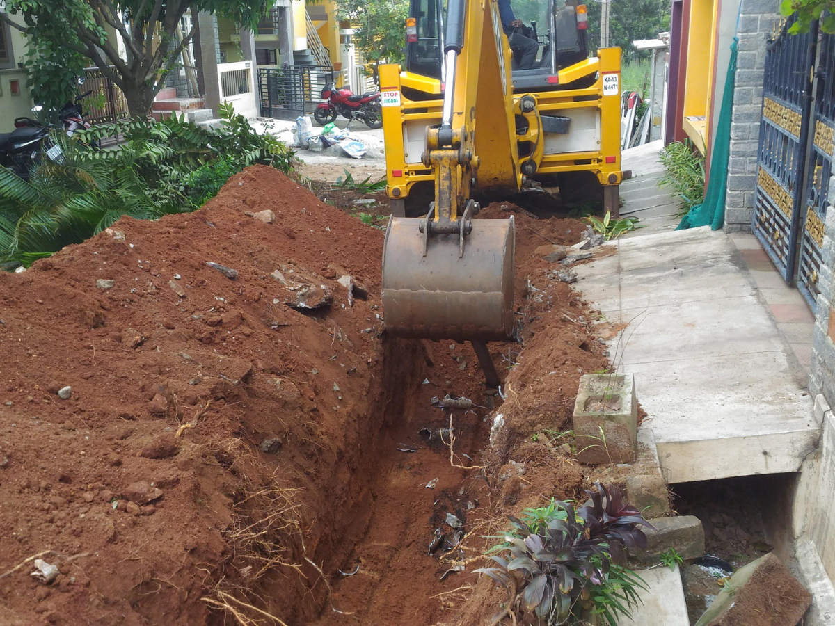 Yellow excavator digging a trench beside a sidewalk, exposing pipes. Yellow excavator digging a trench beside a sidewalk, exposing pipes.