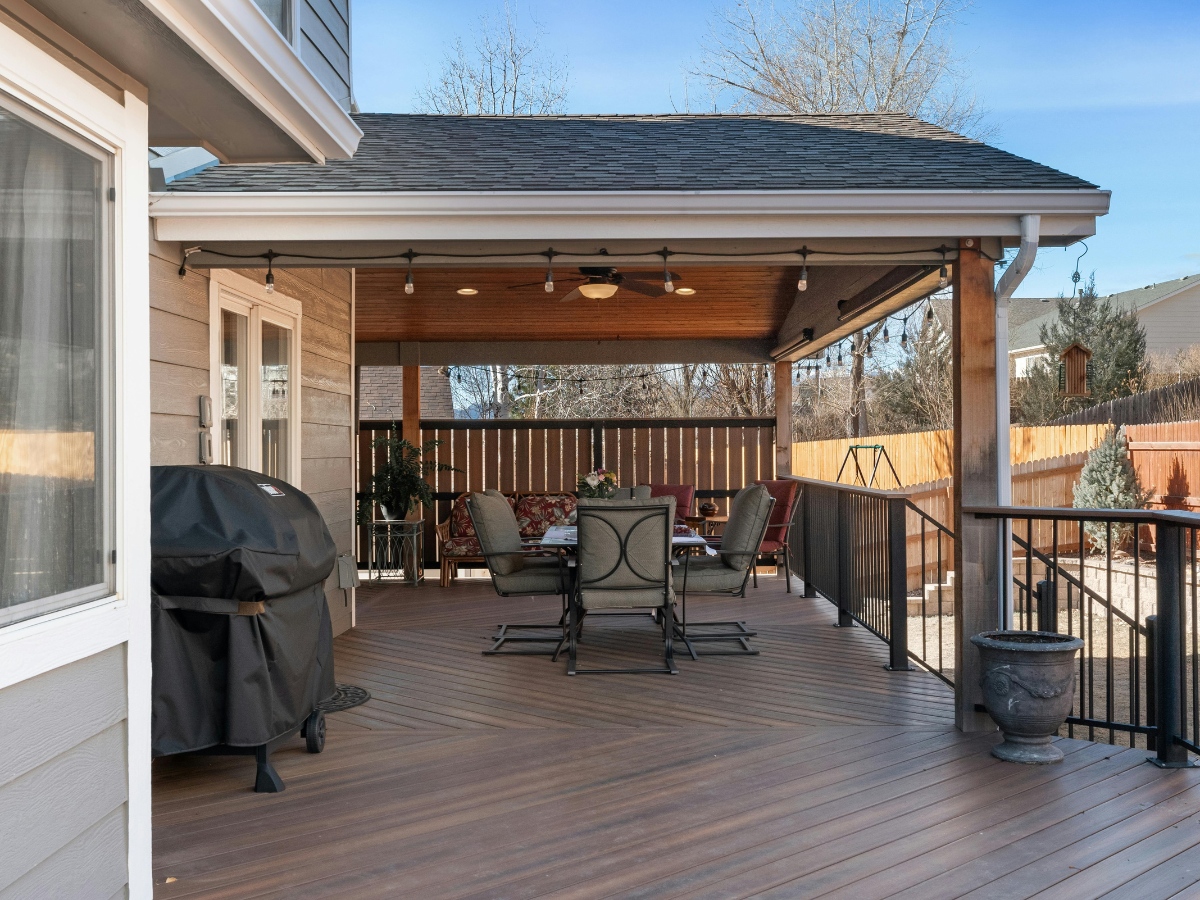 Covered wooden deck with outdoor dining set, grill, and a view of a fenced backyard.