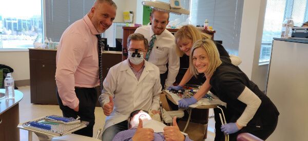 A group of people are standing around a man in a dental chair giving a thumbs up.