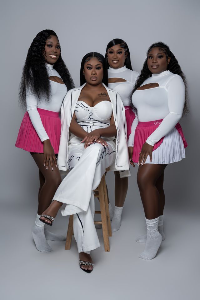 Four women in white and pink outfits posing together; studio background.