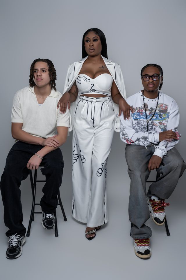 Three people posing: woman in white suit, two others seated. Gray backdrop.