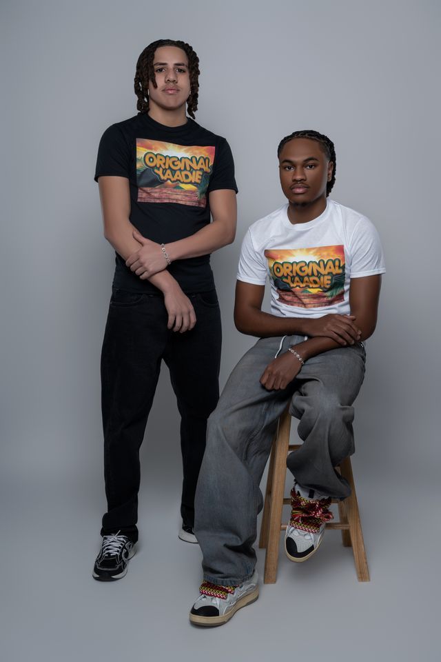 Two people in t-shirts with an image on the front, pose for a photo. One sits on a stool.