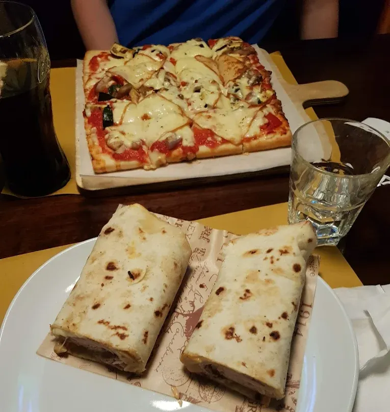 Pizza quadrata su un tagliere di legno, piadine su un piatto, un bicchiere d'acqua e un bicchiere di soda su un tavolo.