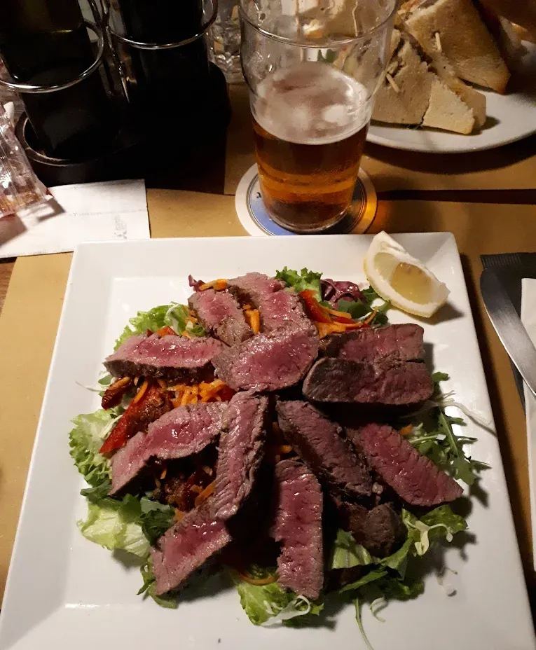 Insalata di bistecca con una fetta di limone su un piatto bianco, accompagnata da un bicchiere di birra.