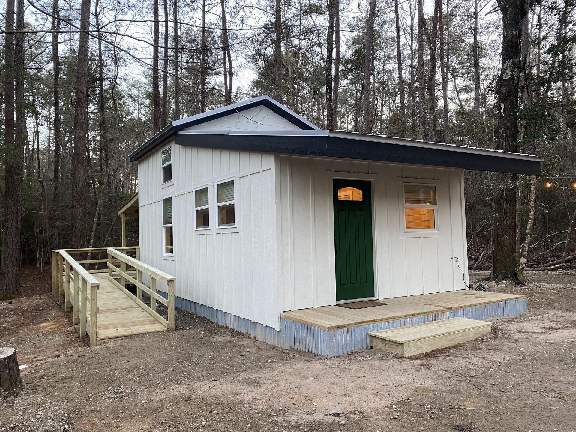 A small white house with a green door and a ramp in the woods.