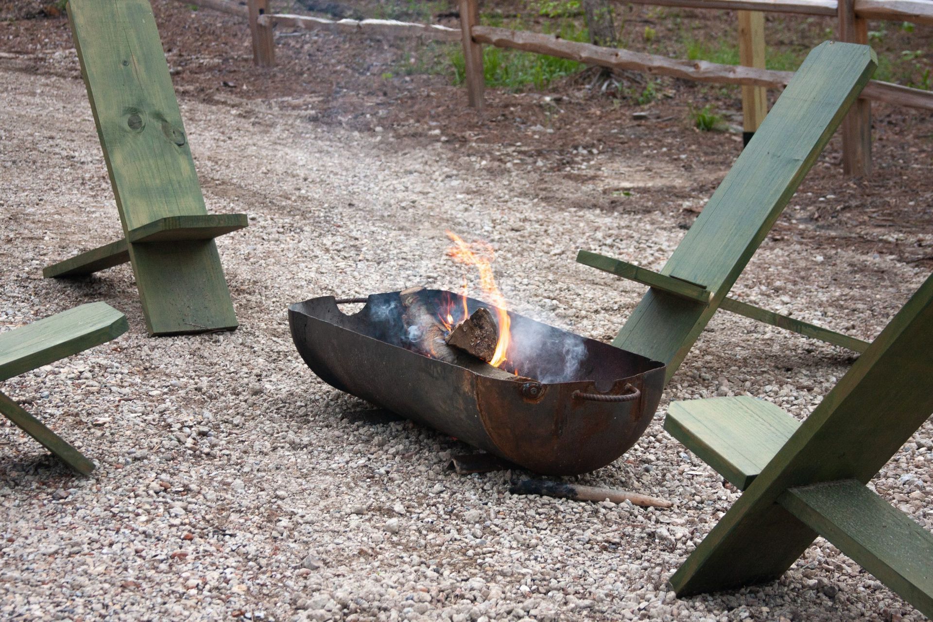 A fire pit is surrounded by wooden chairs in a gravel area.