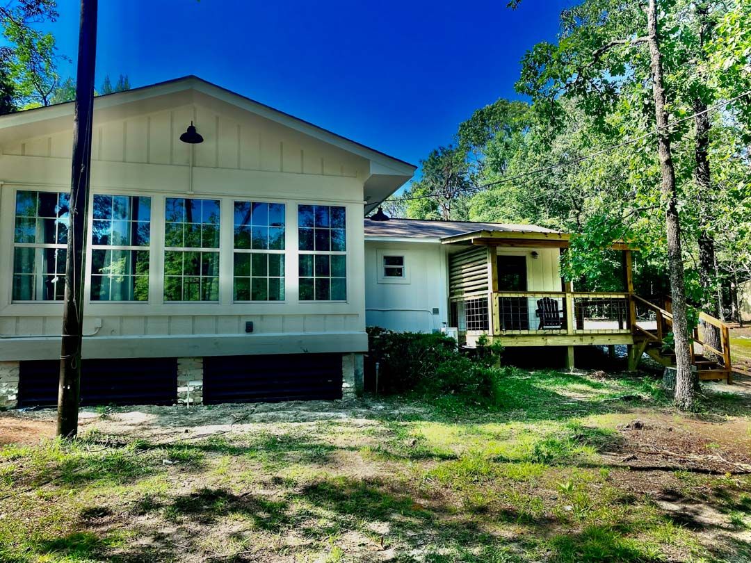 A white house with a large porch and lots of windows is surrounded by trees.