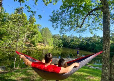 A man and a woman are laying in a hammock near a lake.