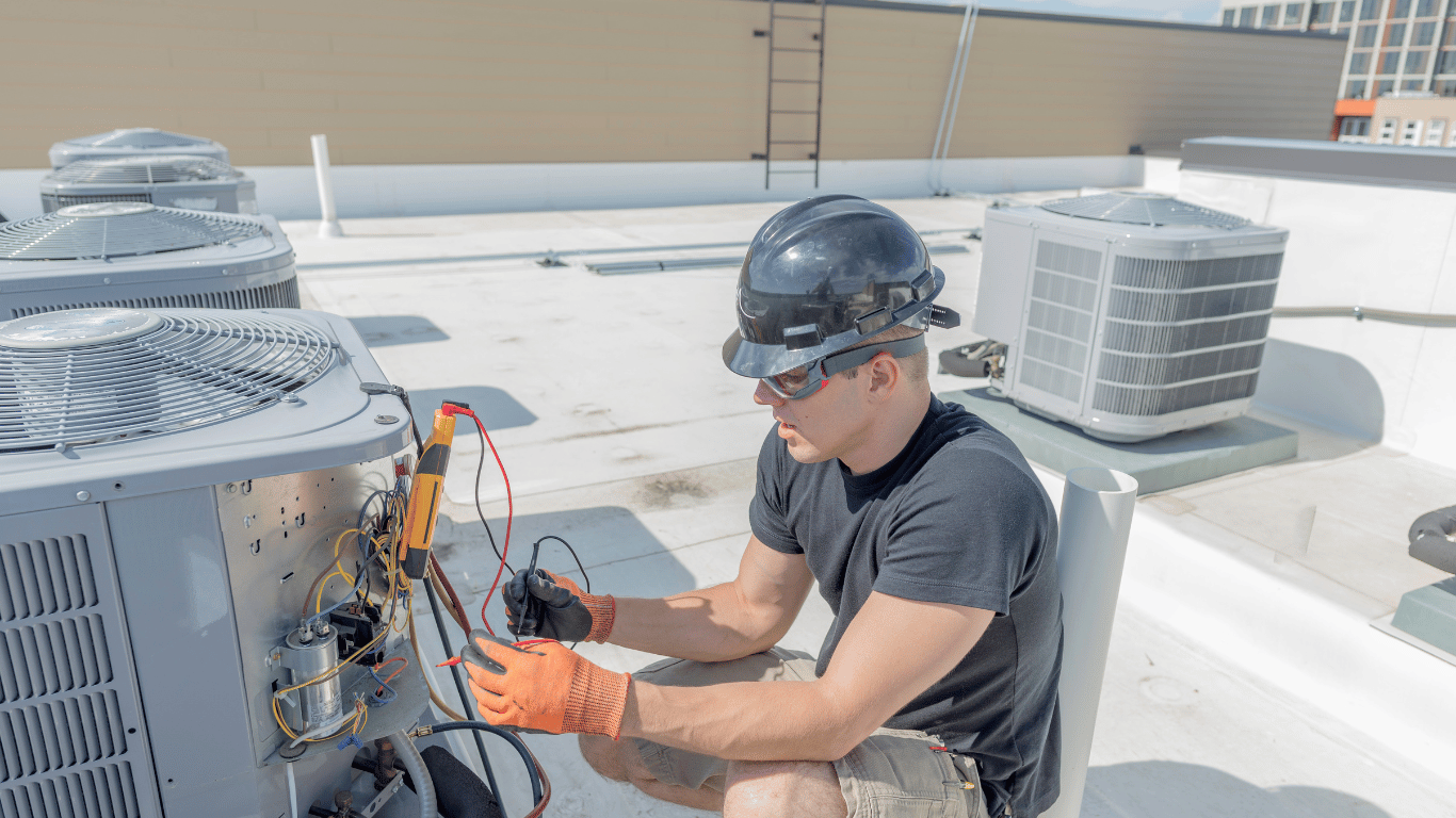 A man is working on an air conditioner on the roof of a building, conducting commercial HVAC repair