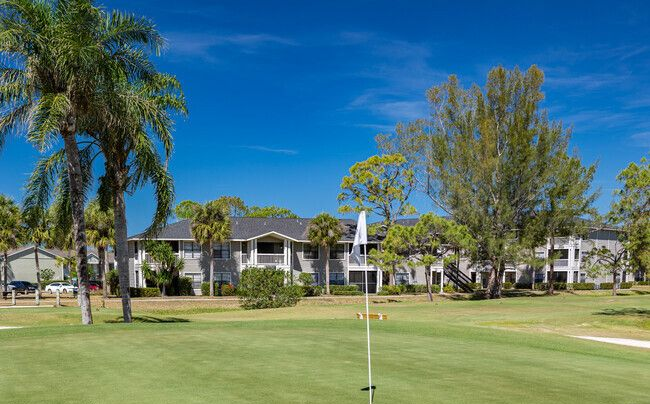 Golf course green with a flag, palm trees, and condos under a clear blue sky.