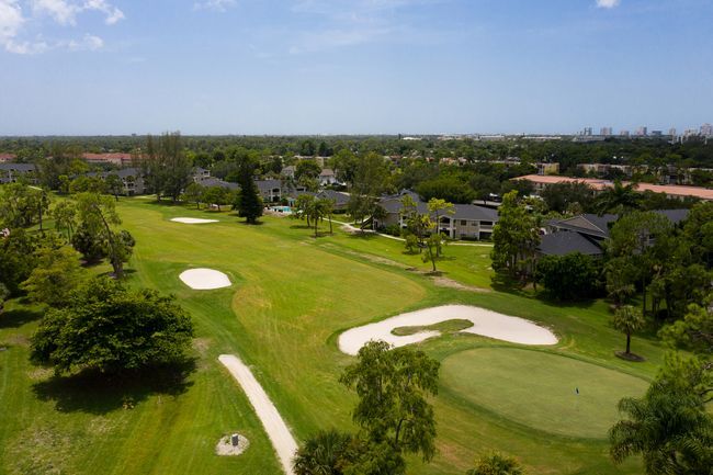 Aerial view of a green golf course with sand traps and trees, buildings in the background under a blue sky.