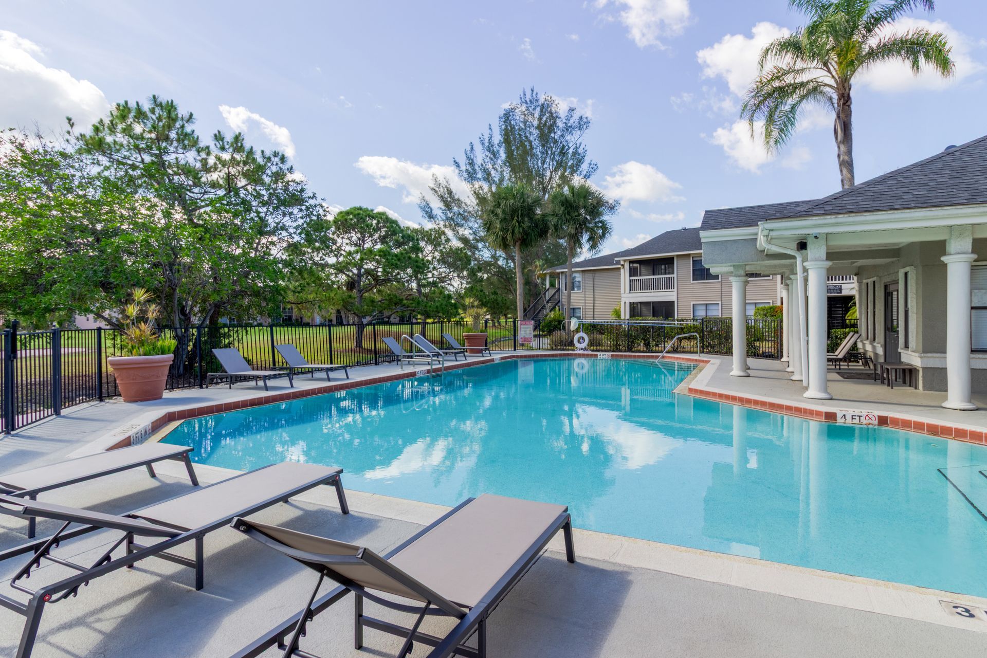 Swimming pool with lounge chairs, trees, and apartment buildings on a sunny day at Belvedere at Quail Run in Naples, FL.