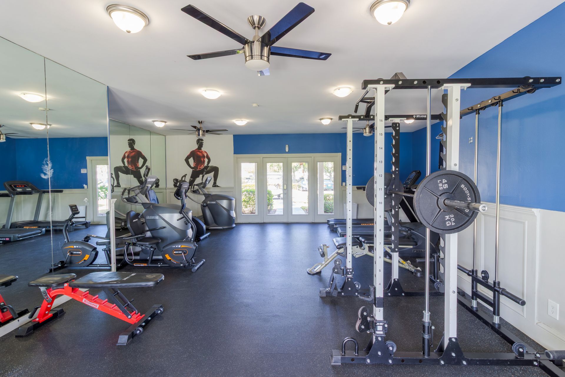 Gym with exercise equipment, including weight machines, treadmills, and a weight bench. Blue walls, mirrors, and ceiling fans at Belvedere at Quail Run in Naples, FL.