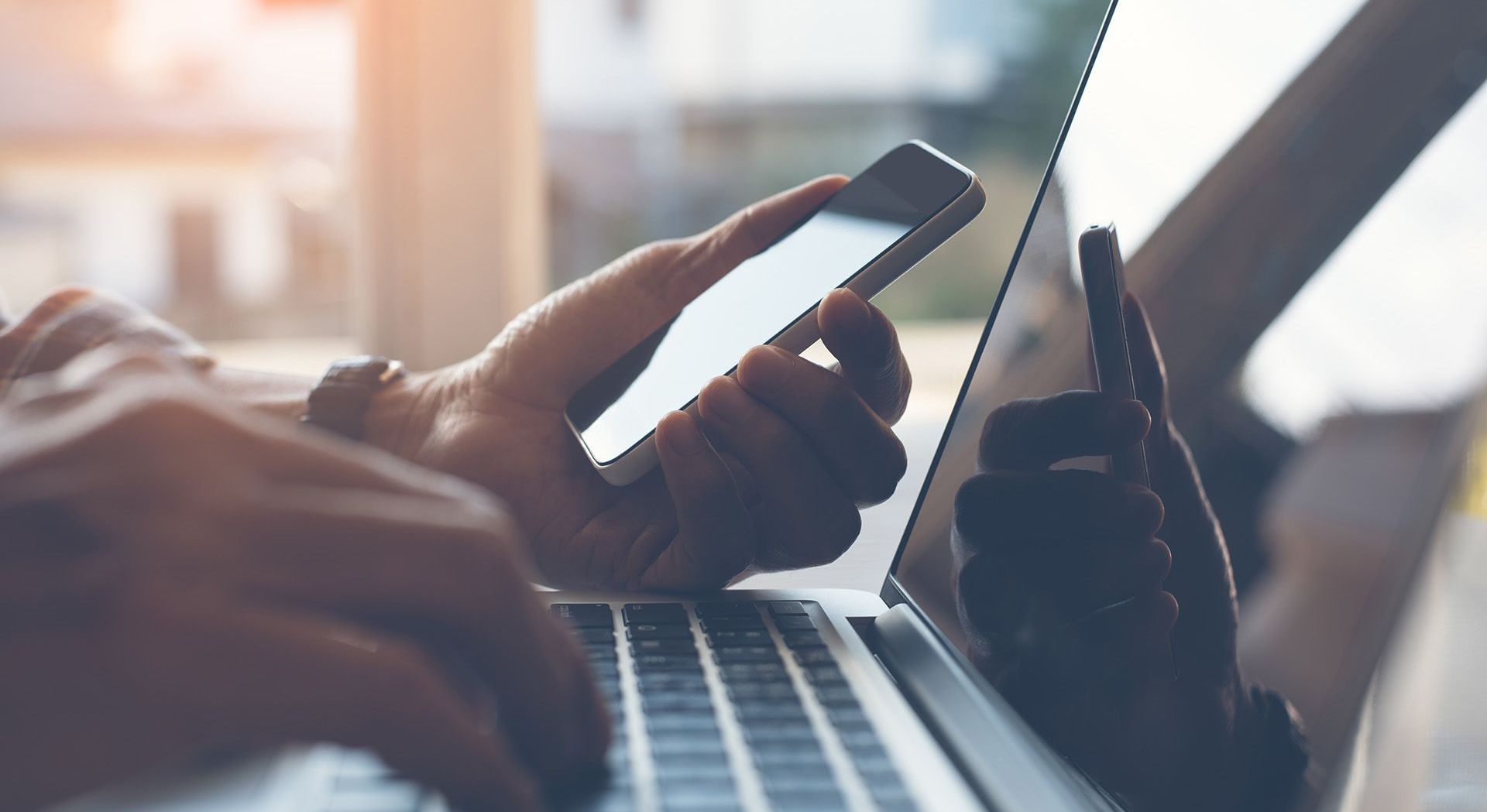 A person is typing on a laptop while holding a cell phone.