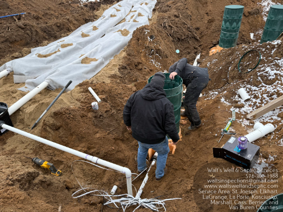 Two workers installing septic system components in a trench. Pipes, tanks, and electrical components are visible.