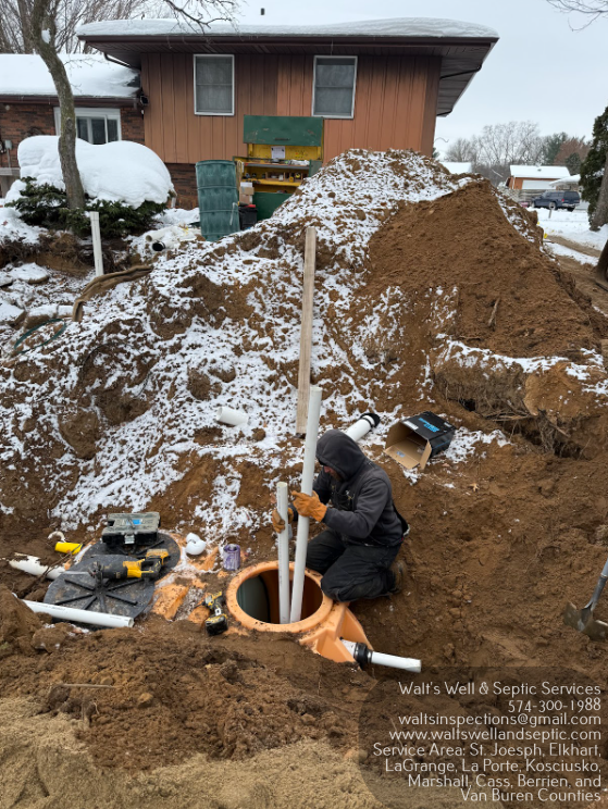 Person installing pipes into a septic system, surrounded by dirt and snow. House in background.