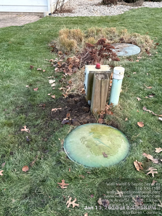 Utility box and white pipe cover in a grassy yard beside a stone ring and bare shrubs
