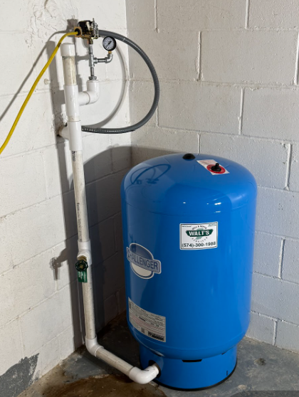 Blue water pressure tank with white piping and pressure gauge against a concrete block wall.