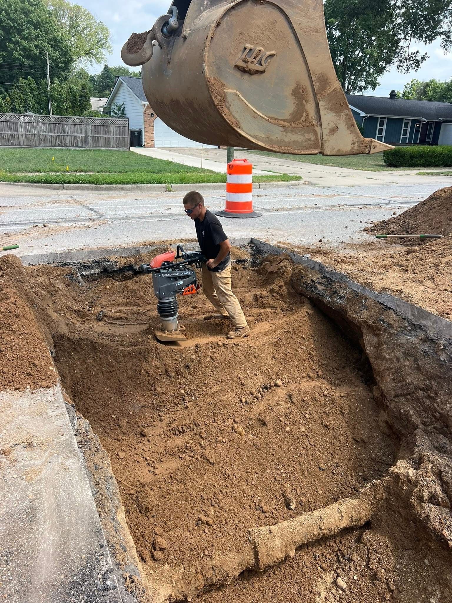 Worker compacting soil in a road repair excavation; excavator bucket overhead.