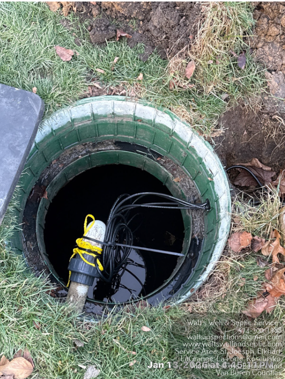 Open septic tank in grass; dark interior with electrical wires and yellow-taped connector.