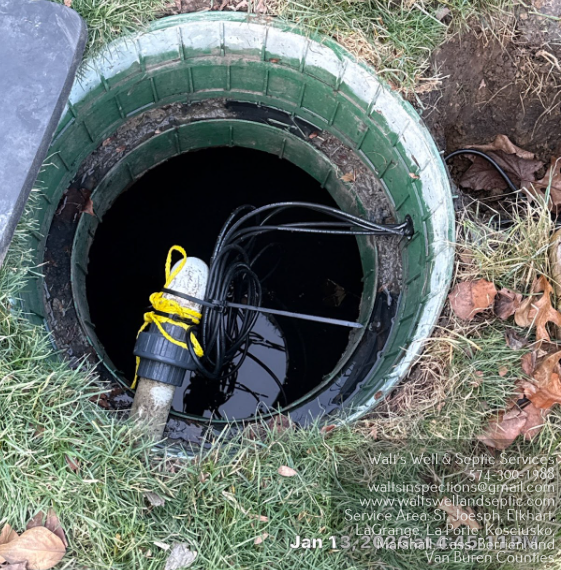 Open manhole with a yellow-and-black cable device and wires inside, surrounded by grass.