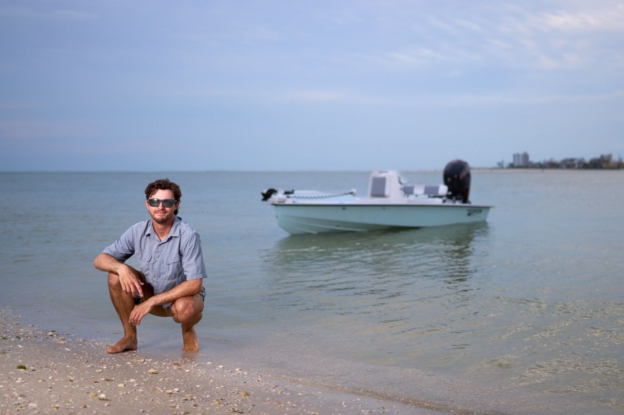 A man is squatting on the beach next to a boat.
