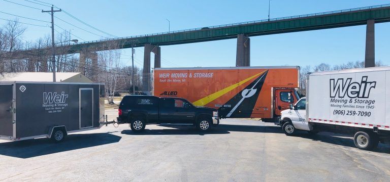 Three moving trucks are parked in a parking lot under a bridge.