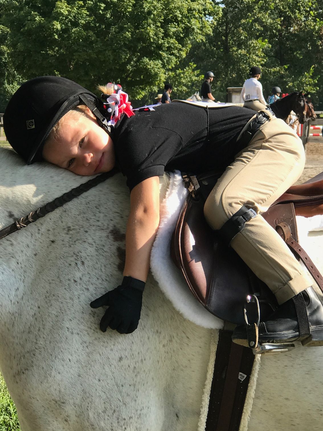 Young rider asleep on a white horse; wearing a black helmet, shirt, tan pants, and black gloves; outdoor setting.
