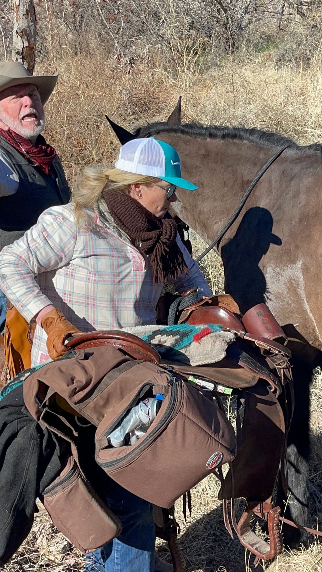 Woman in Western attire adjusting a saddlebag on a horse, man in background. Outdoors.