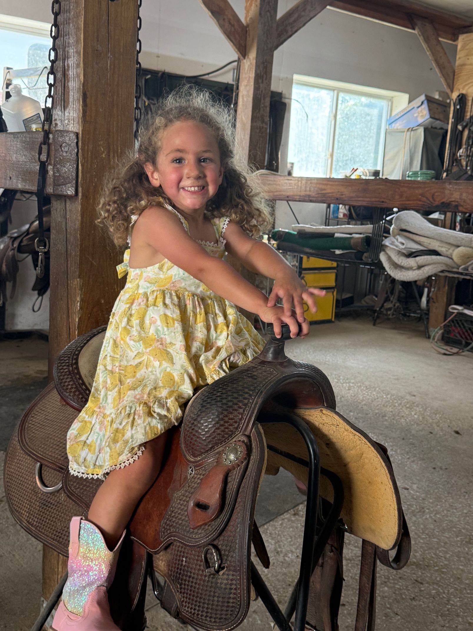 Young girl in floral dress smiles while sitting on a saddle in a barn.