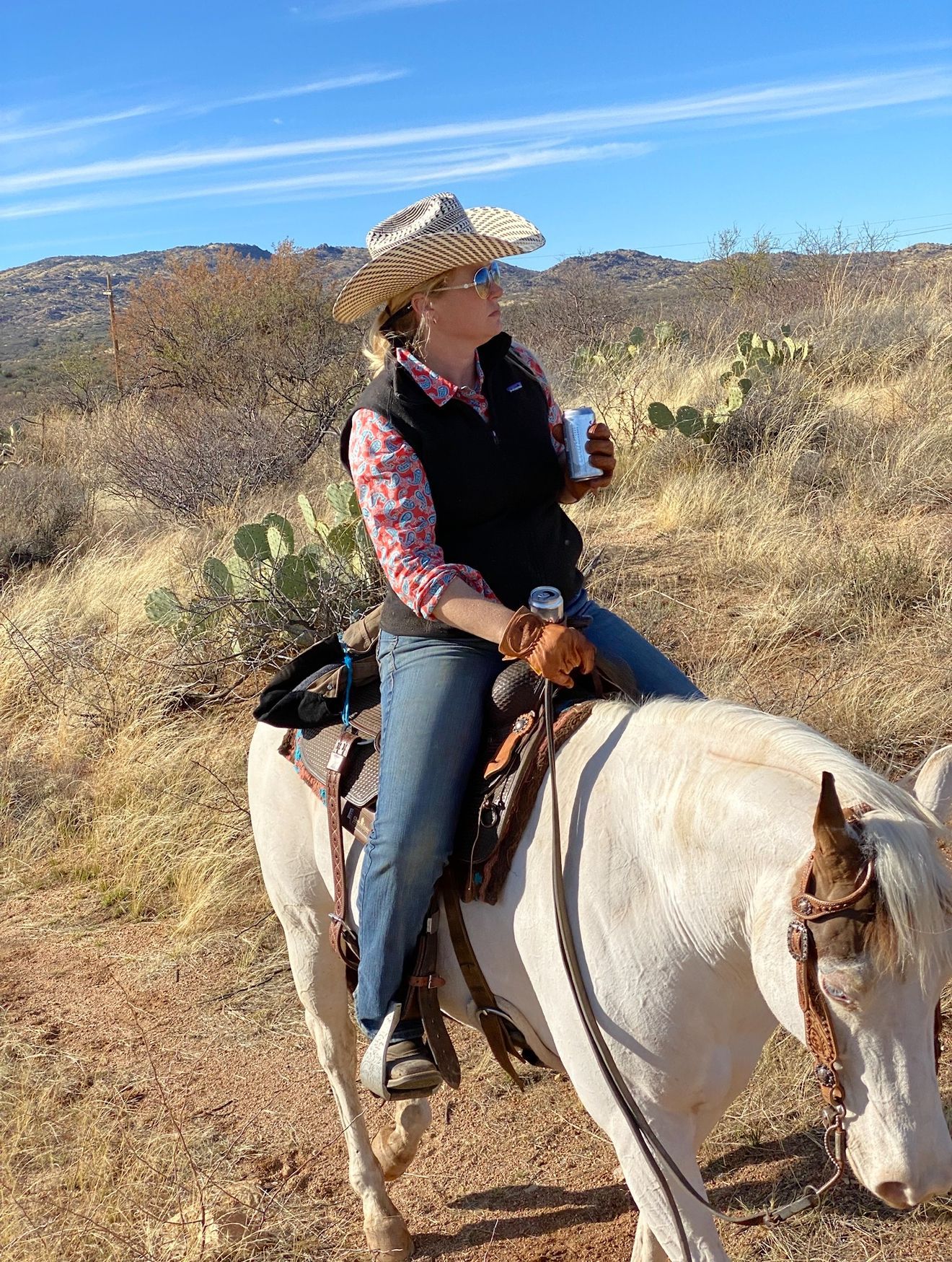 Woman in cowboy hat on a white horse, holding a can, looking up at a blue sky in a desert setting.
