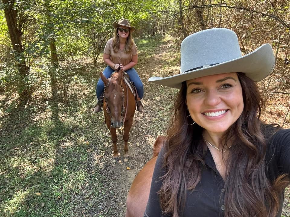 Woman in a cowboy hat smiles, taking a selfie with a friend riding a horse on a trail.
