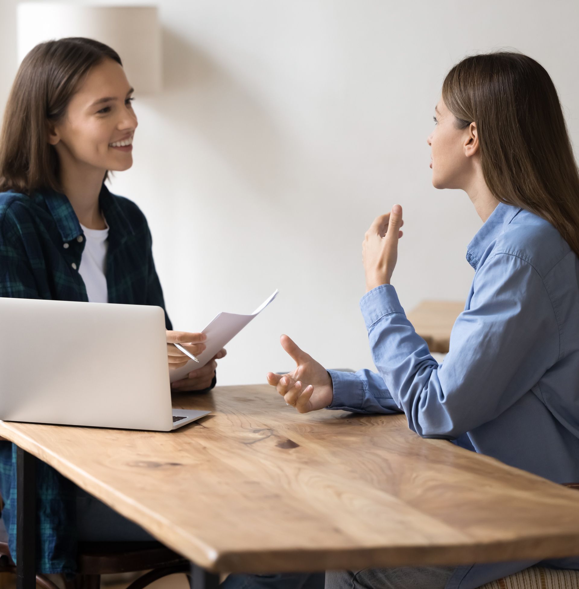Two women at a wooden table; one holds papers, other gestures while speaking.