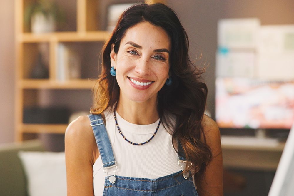 Woman with a warm smile wearing denim overalls and a white top, standing in a room with a bookshelf.