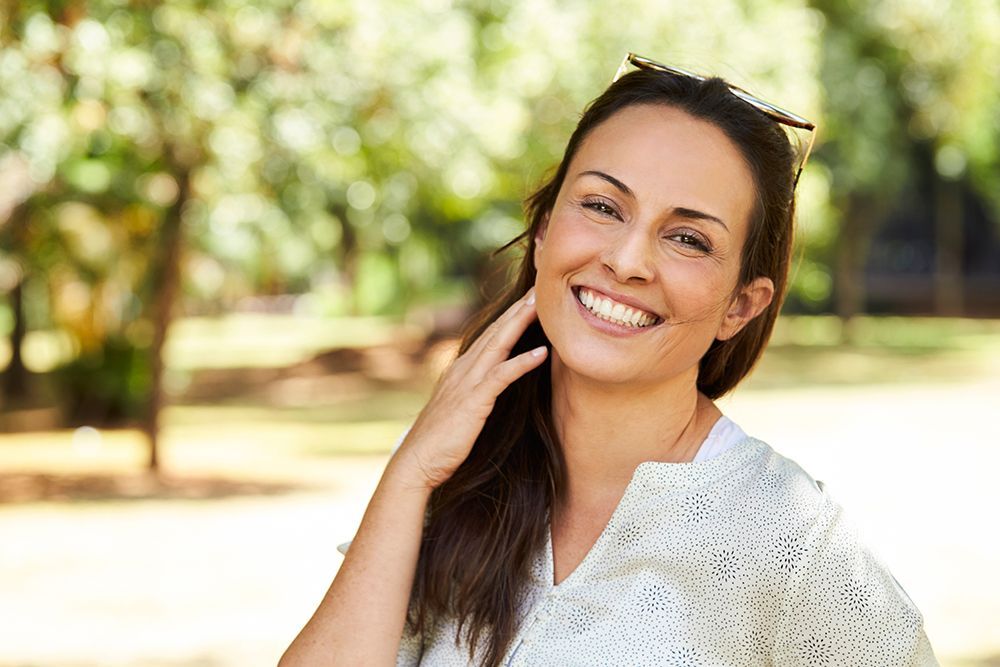 Woman smiling outdoors, hand to her neck, wearing a white shirt; trees in background.