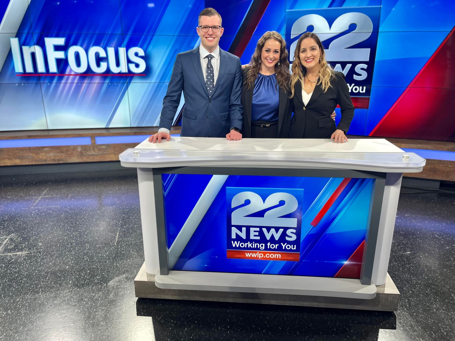 Three news anchors stand behind a desk in a studio. The background has a blue graphic with the station's logo.