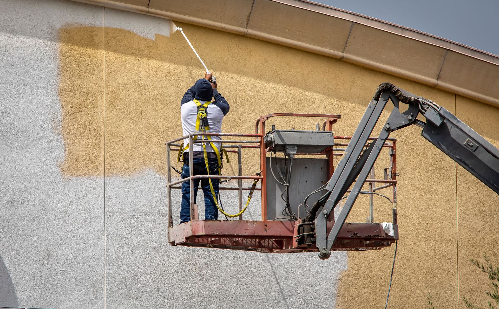 A worker in a yellow shirt hangs from ropes, using a roller to paint a white stripe on a blue building wall.