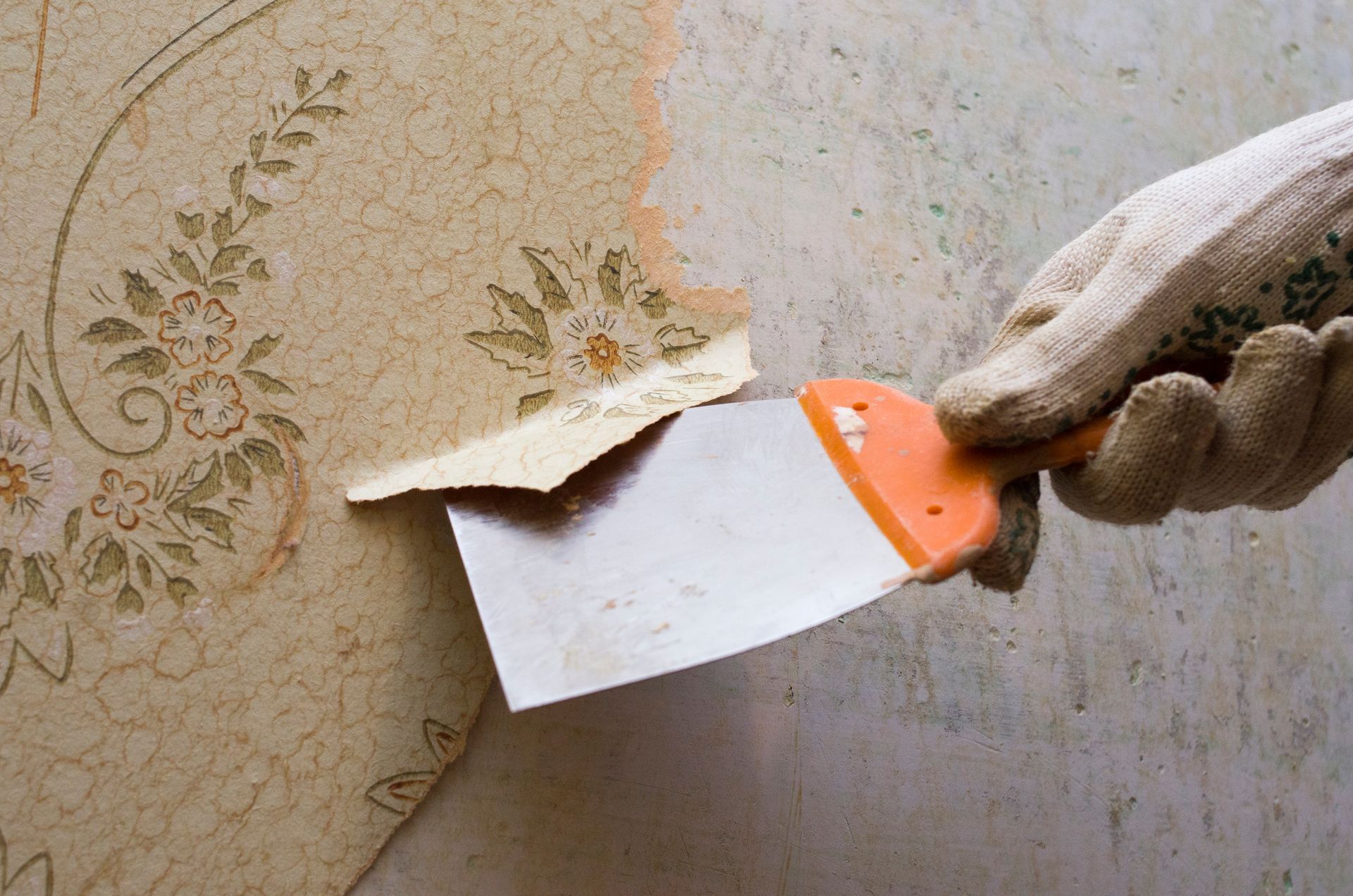 A gloved hand uses a putty knife to scrape old, patterned floral wallpaper off a wall.